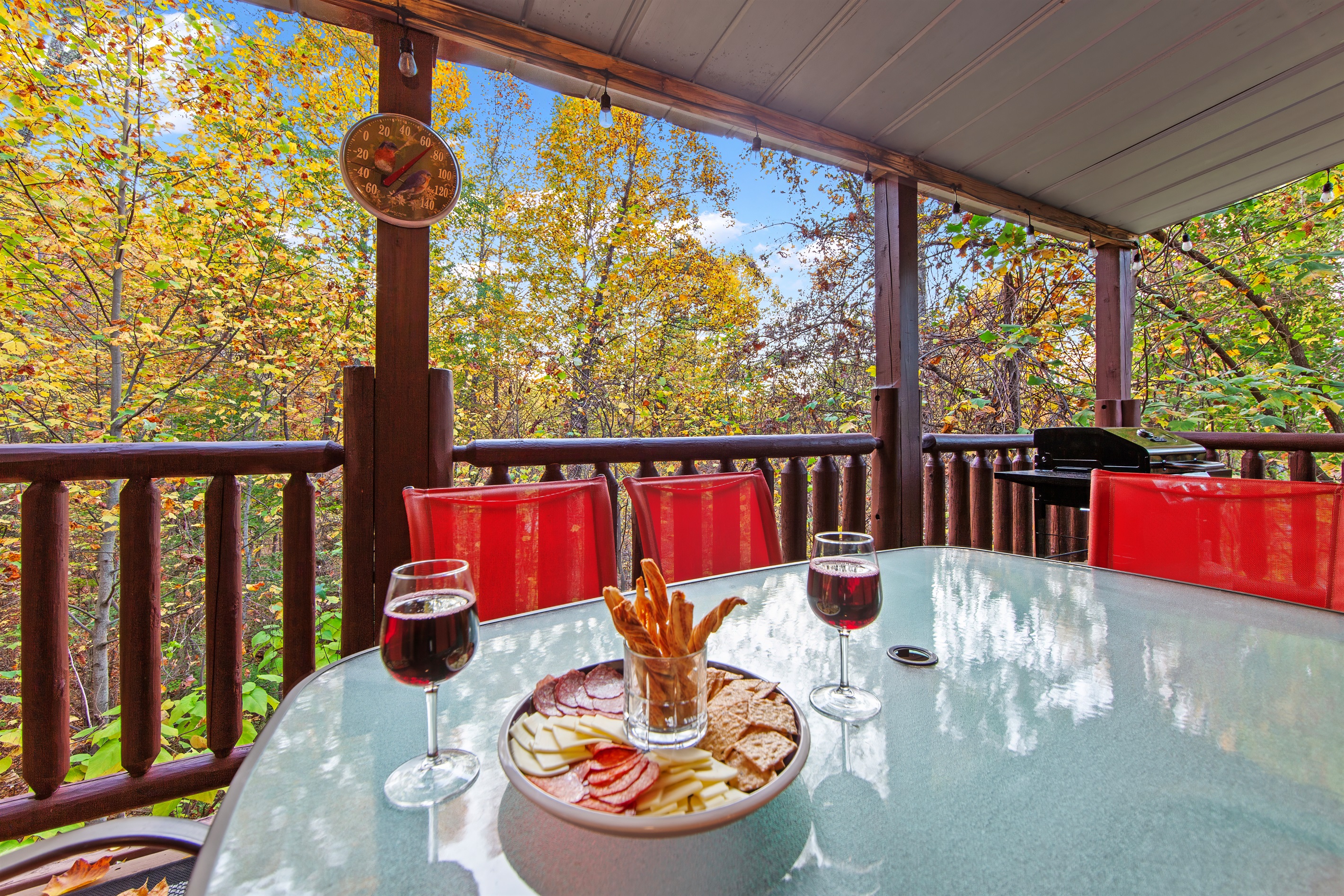A delightful deck with a glass dining table, vibrant red chairs, and a grill under a covered roof offers a comfortable and scenic spot to dine or unwind in the heart of nature.