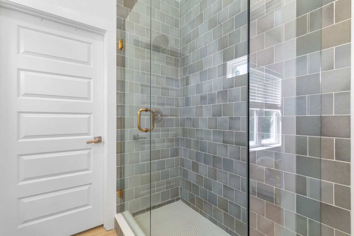 The shower takes up a full corner, tiled in blue and grey with a small window letting the daylight in. Brass hardware on the glass, honeycomb underfoot, and room enough to actually move around in there.