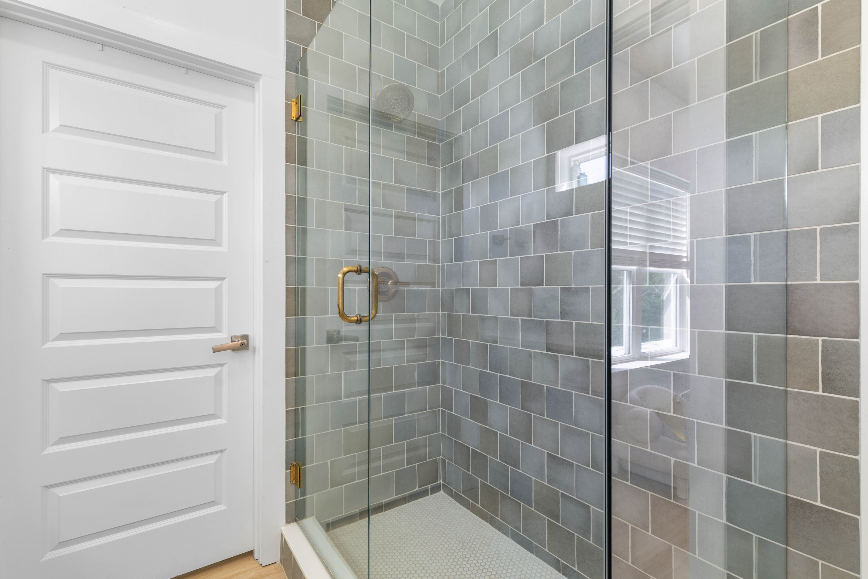 The shower takes up a full corner, tiled in blue and grey with a small window letting the daylight in. Brass hardware on the glass, honeycomb underfoot, and room enough to actually move around in there.