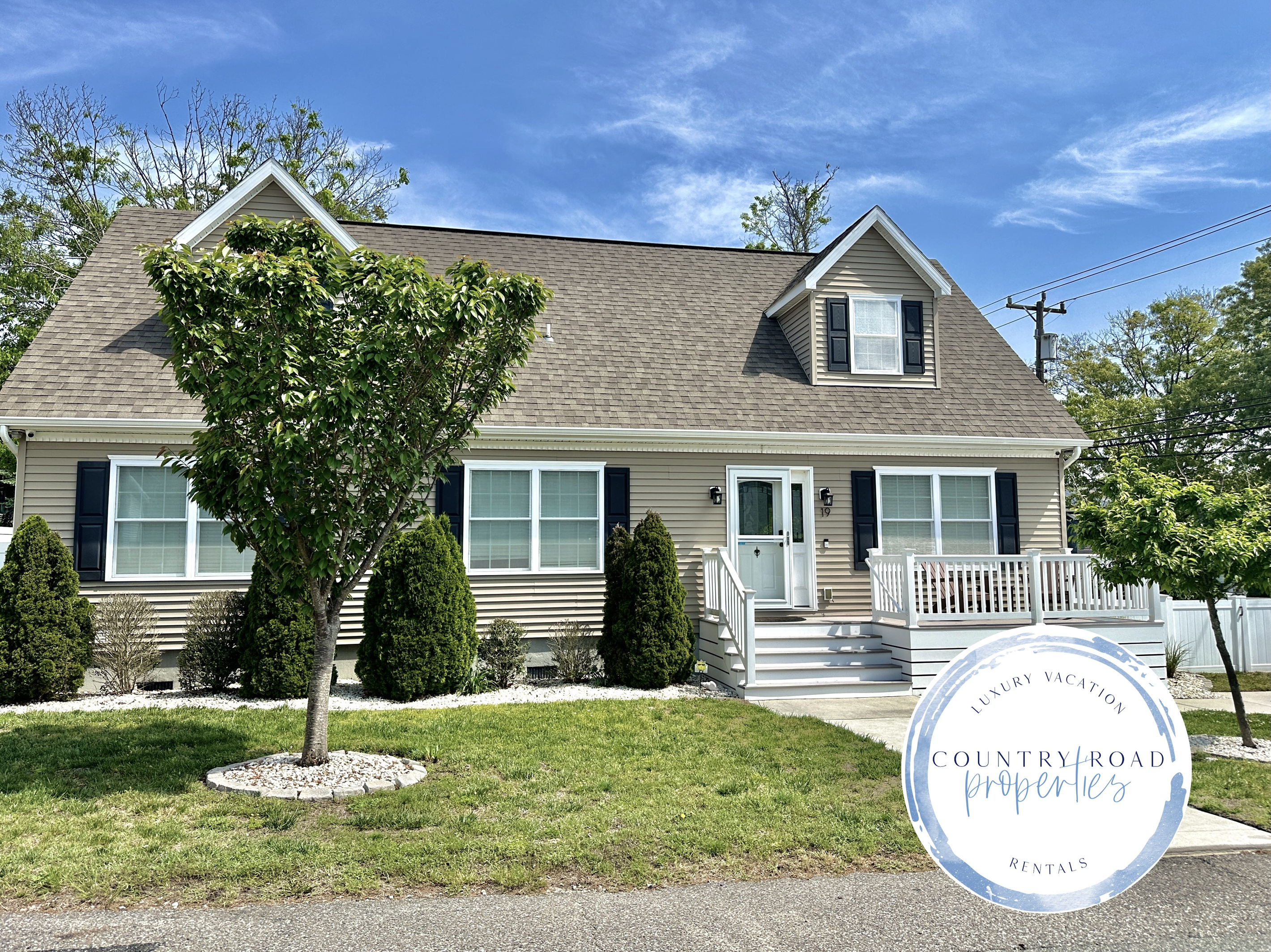 This welcoming home features a clean tan exterior with a small front porch and white railings. The property includes a green lawn, several evergreen shrubs, and a concrete walkway leading to the front door.