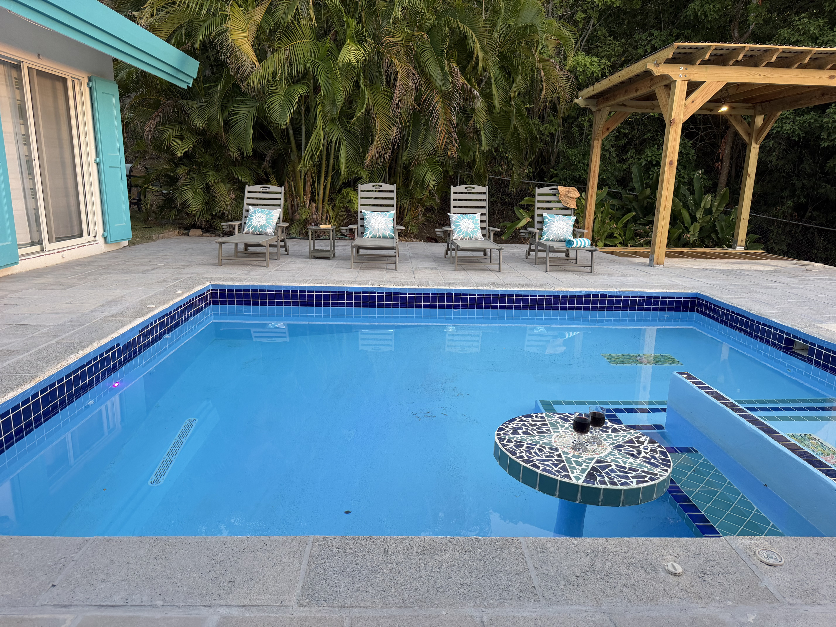 A tiled, circular swim-up table set inside the water, perfect for resting drinks or enjoying evening cocktails. A wooden gazebo with overhead lighting, offering shelter and cozy evening illumination beside the deck