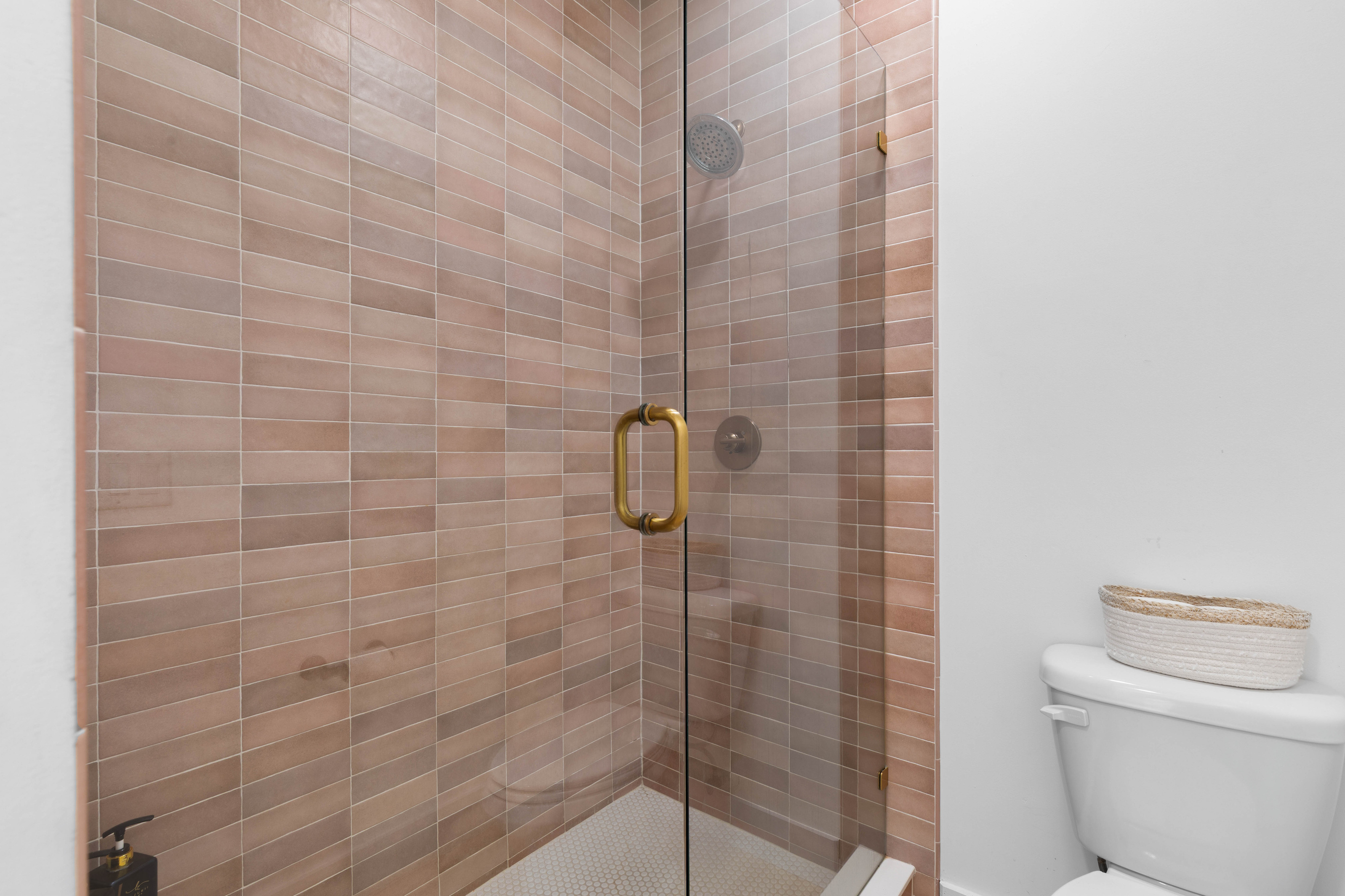 The walk-in shower runs floor to ceiling in warm clay tile, with a brass handle on the glass. After a morning out on the sand this is the first stop, and the honeycomb floor keeps footing sure underneath.