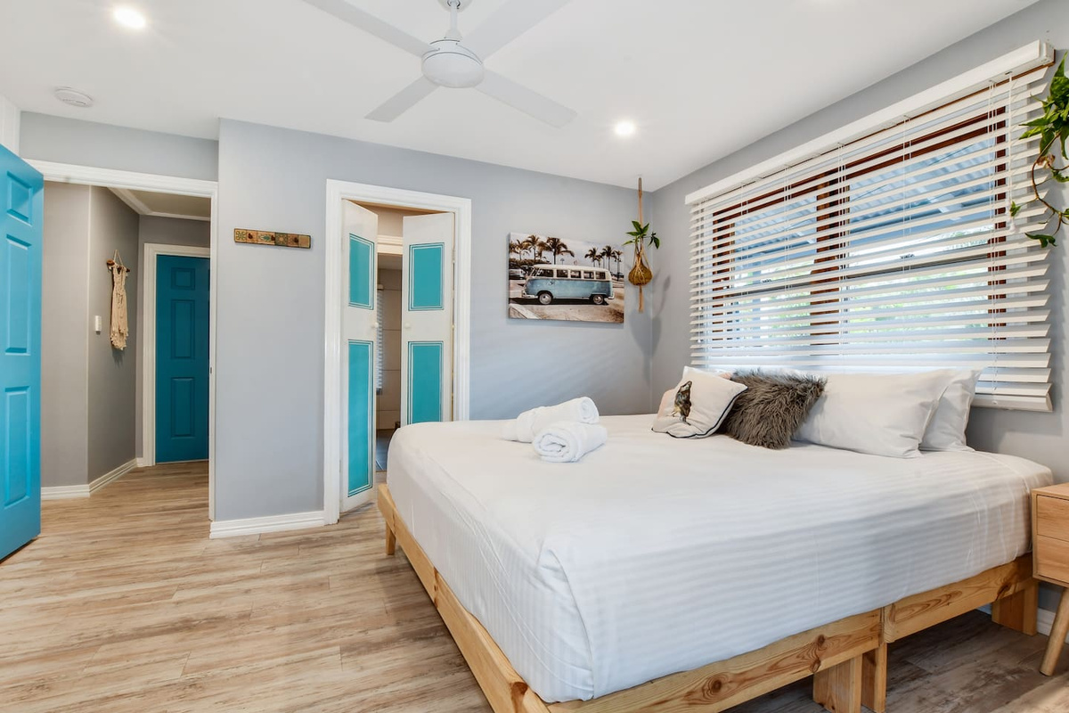 Aqua doors, timber blinds and light coastal finishes give this queen bedroom at Aaron's Beachside Boat House a distinctive beach-house personality.
