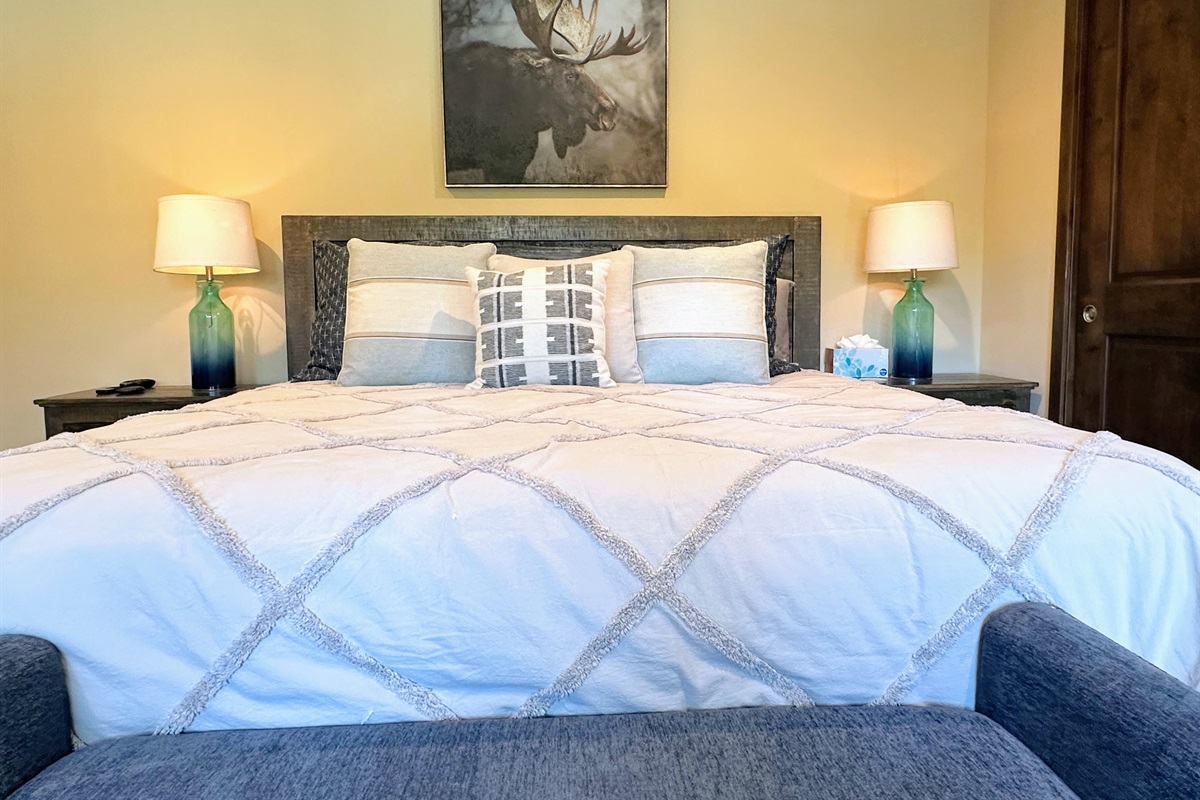 Style meets serenity with a king bed framed by matching side tables and blue glass lamps, a cozy blue bench at the foot, and striking nature-inspired art that grounds the room in calm, natural beauty.