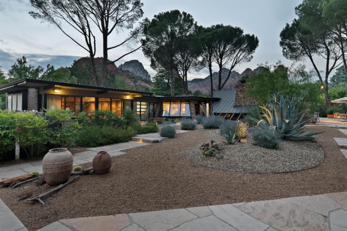 Spectacular dusk view of this mid-century sanctuary, nestled among mature pines and framed by Sedona’s iconic red rock formations — where serene desert landscaping meets timeless architectural design.