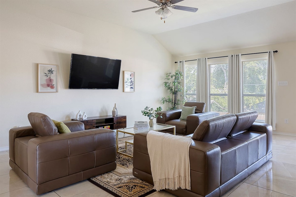 Welcome to this elegant living room with plush leather sofas and a large wall-mounted TV, bathed in natural light.