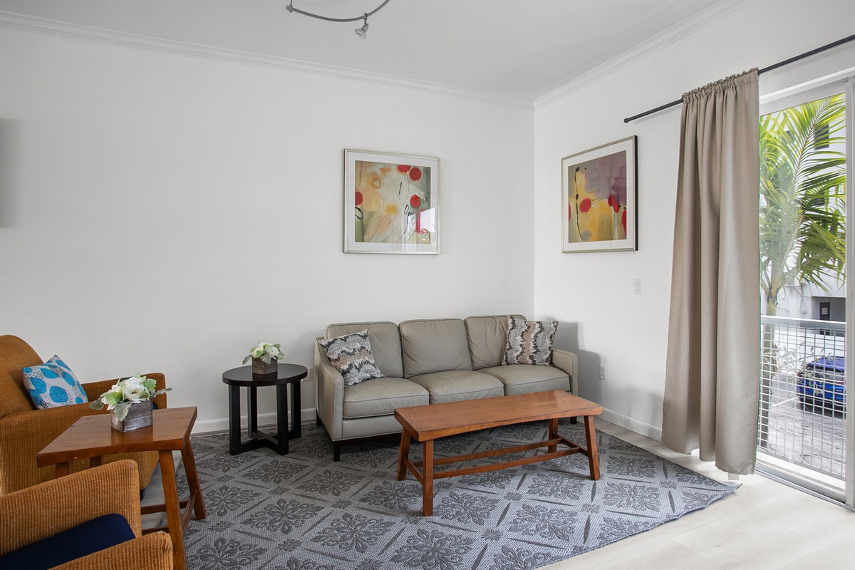 Living room with leather sofa, accent chairs, and a 50-inch flat screen TV. Sliding glass doors open to a Juliet balcony for sea breeze.