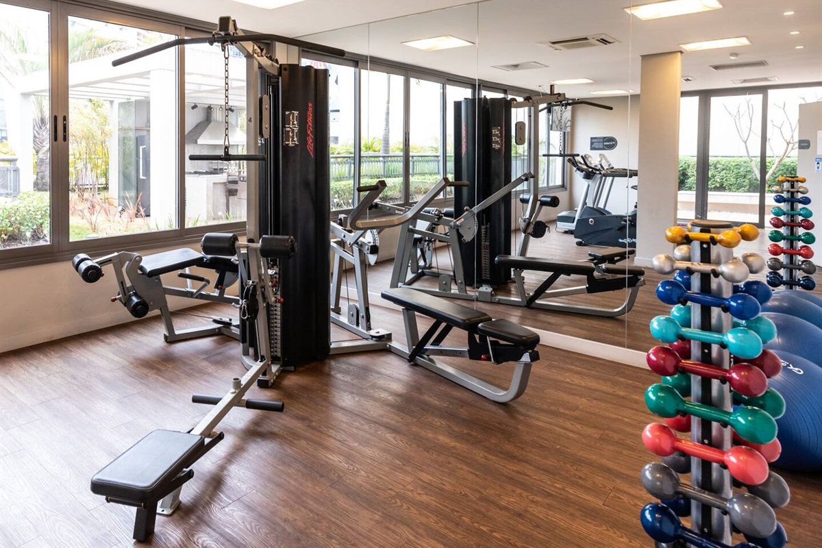 Espaço de musculação com pesos livres, estação multiuso e vista iluminada.