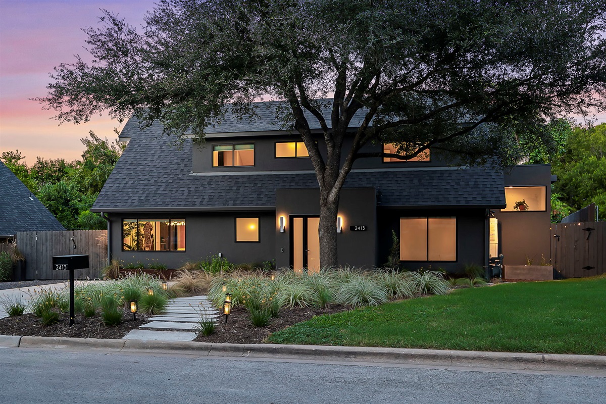 A stunning modern home with a dark gray facade, a prominent A-frame roof, and a minimalist landscape design with a stone pathway, all illuminated at dusk to create an inviting and sophisticated curb appeal.