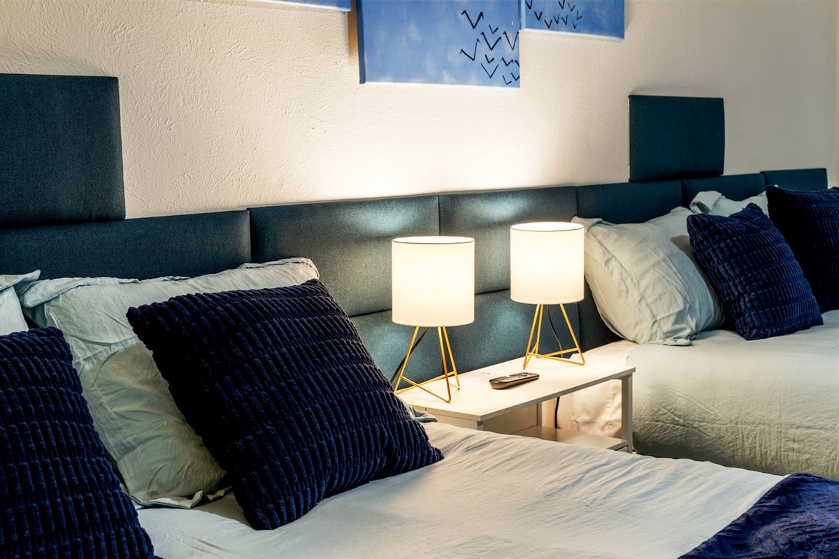 Spacious bedroom with two plush queen beds, blackout curtains, and a smart TV—ideal for families or groups to unwind in comfort.