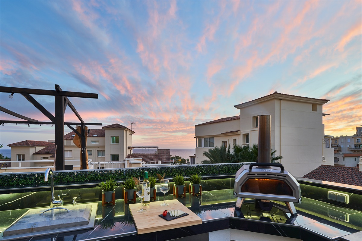 Fire up the built-in barbecue or the wood-fired pizza oven and enjoy cooking with a view. The fully equipped outdoor kitchen features a sink, fridge, and prep space with subtle lighting for evening meals. Dine under the open sky at the outdoor table, surro