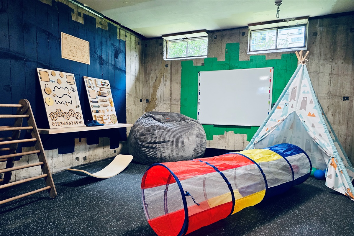 Teepee, climbers, sensory boards, and creative tools fill this industrial-chic play zone. Toys rotate by age, and a ceiling-mounted sensory swing is stored in the bench for added fun.
