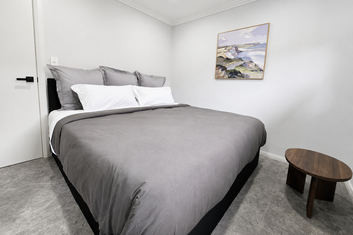 This bedroom includes a King bed with grey and white bedding, tiled flooring, a side table and framed artwork.