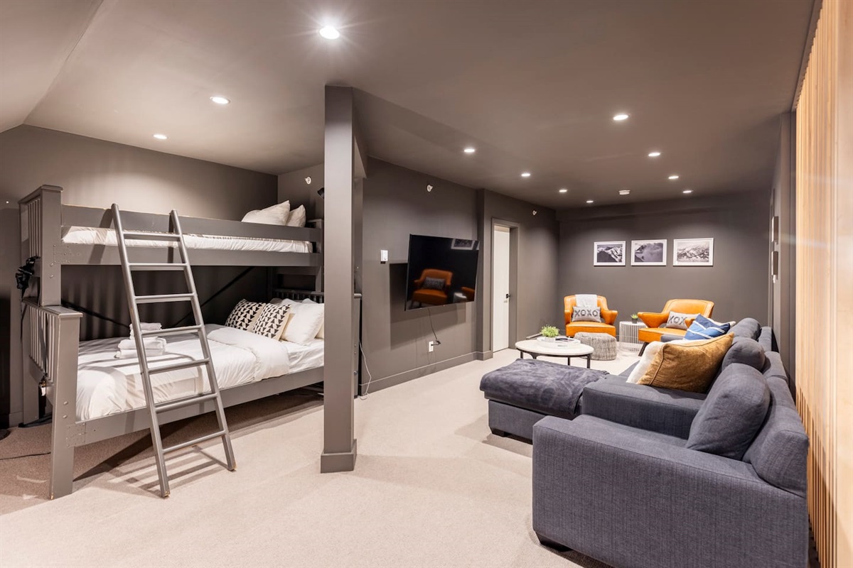 Large upstairs media room has a 65" TV, single  over queen bunk beds and sectional couch and chairs for lounging. The full bathroom is through the door to the right of the TV.