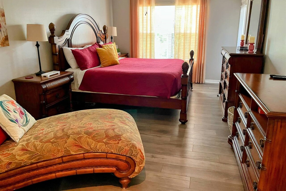 Spacious bedroom featuring a large bed, chaise lounge, wood-look flooring, dresser storage, and warm natural light.