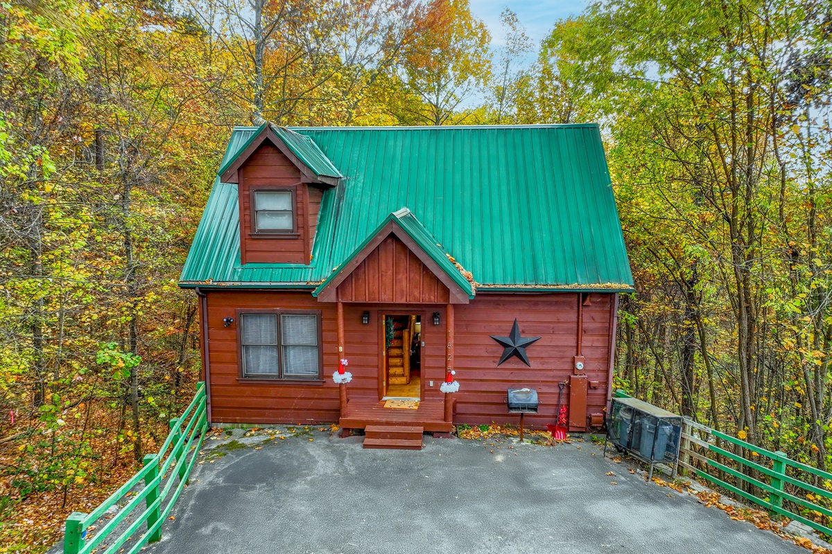 This inviting cabin showcases a green roof and warm wood tones, enhanced by soft porch lighting and a star accent, nestled among the natural beauty of the forest.