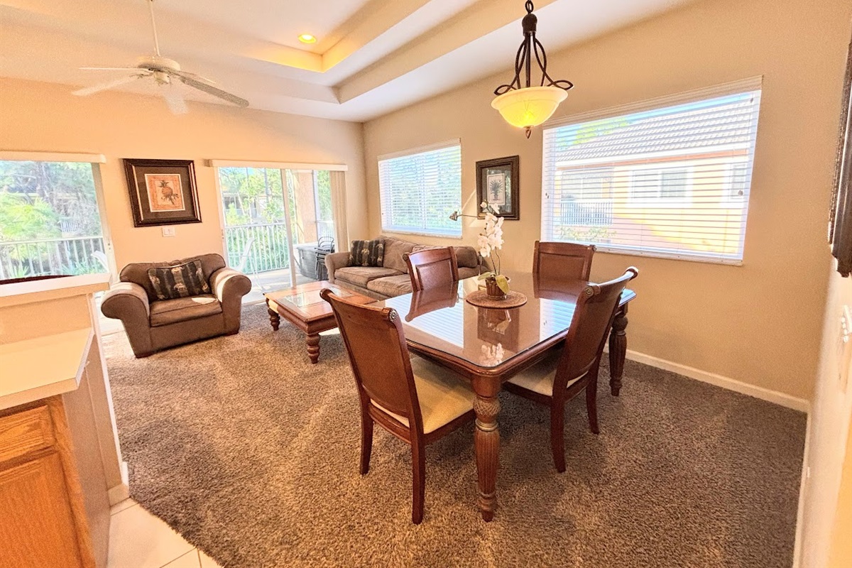 Open dining and living area with comfortable seating, tray ceilings, large windows, and sliding glass doors leading to the private balcony.