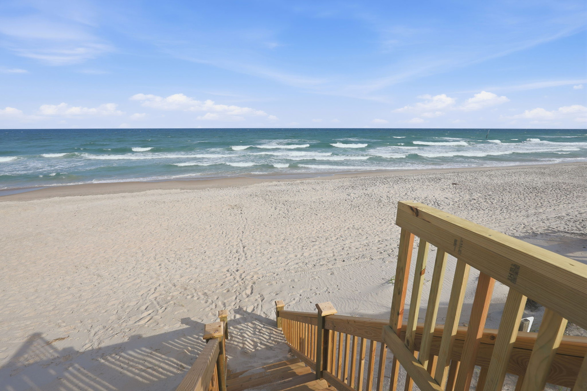 Ground-level beach entry perspective from the stairway, looking out over the wide sandy beach and ocean