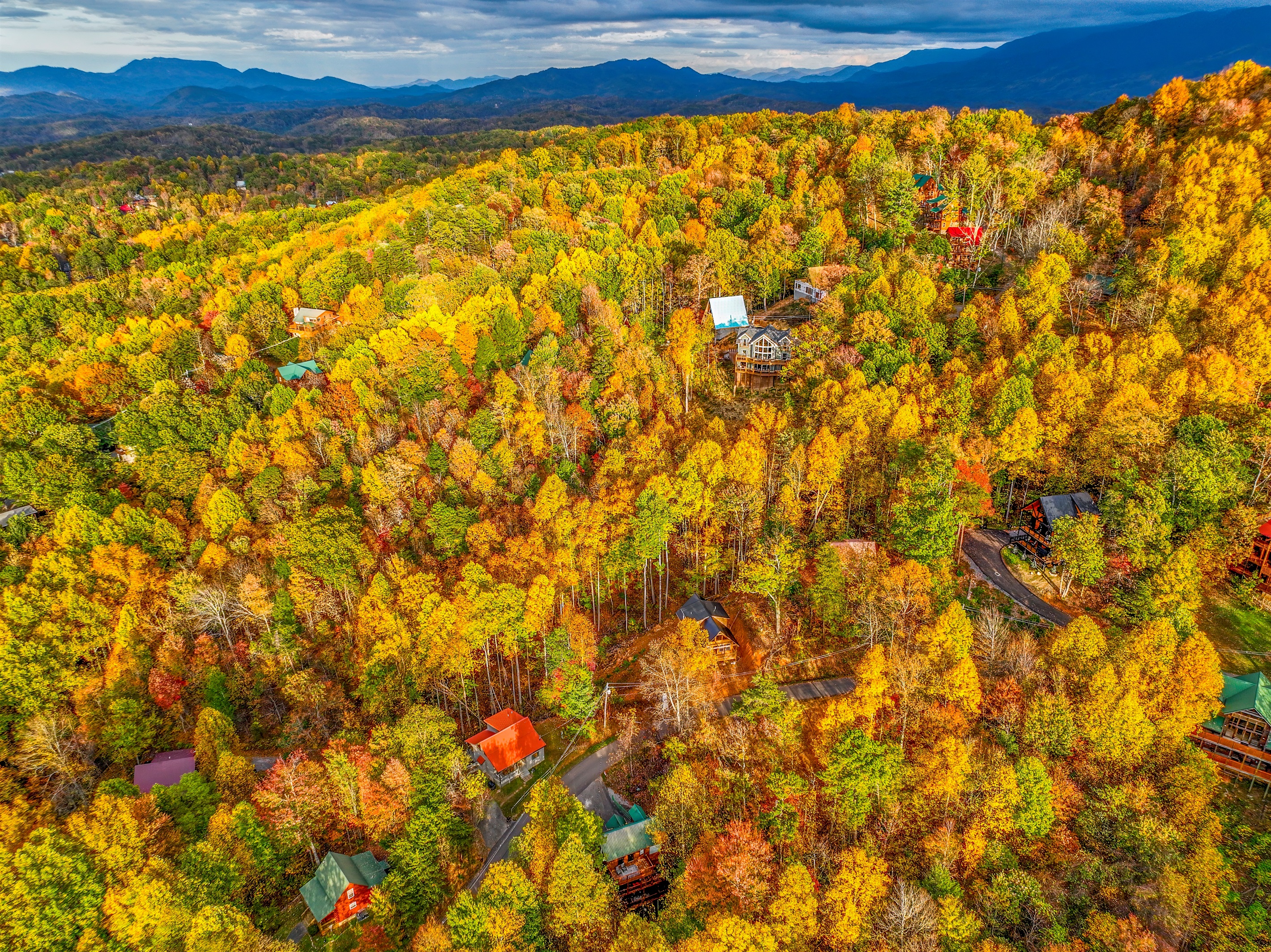 An aerial view of a serene autumn landscape, where vibrant hues of gold, orange, and red blanket the forest. Cozy cabins nestle among the trees, with majestic mountains rising in the distance.
