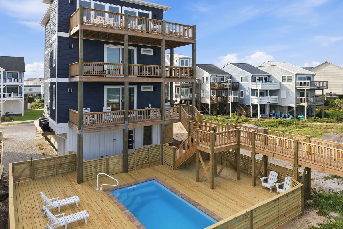 Angled exterior pool-and-home view showing the main house, surrounding decks, and neighboring beach homes, emphasizing the multi-level architecture, private outdoor spaces, and close proximity to the ocean
