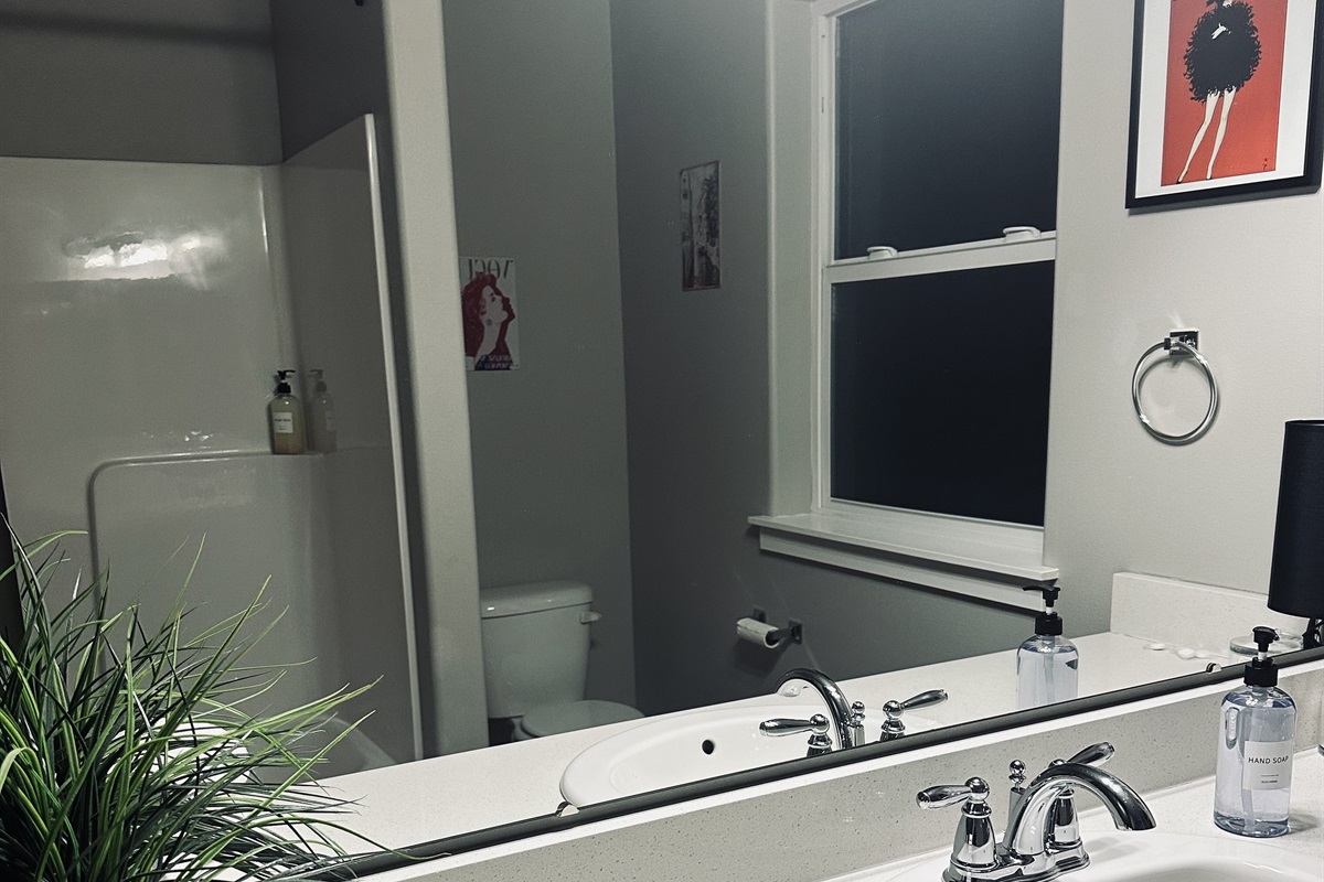 A stylish bathroom features a sleek white countertop with a faucet, a mirror reflecting the space, two bottles of hand soap, a small black lamp, a window with dark surroundings visible outside, a white toilet, and a shower stall, all accented by green foliage in the foreground.