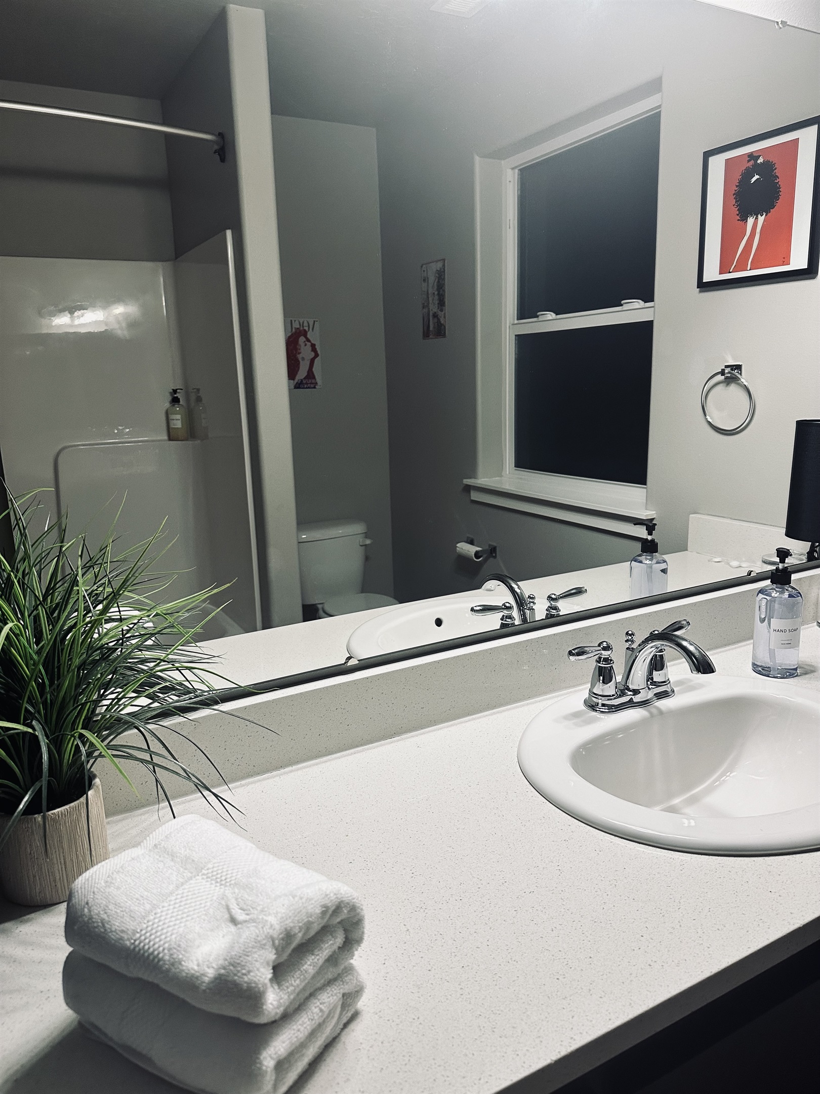 A modern bathroom features a white countertop with two neatly rolled towels, a small potted plant, a sink with chrome faucets, and a large mirror reflecting a window and artwork on the walls, while a shower area is visible in the background.