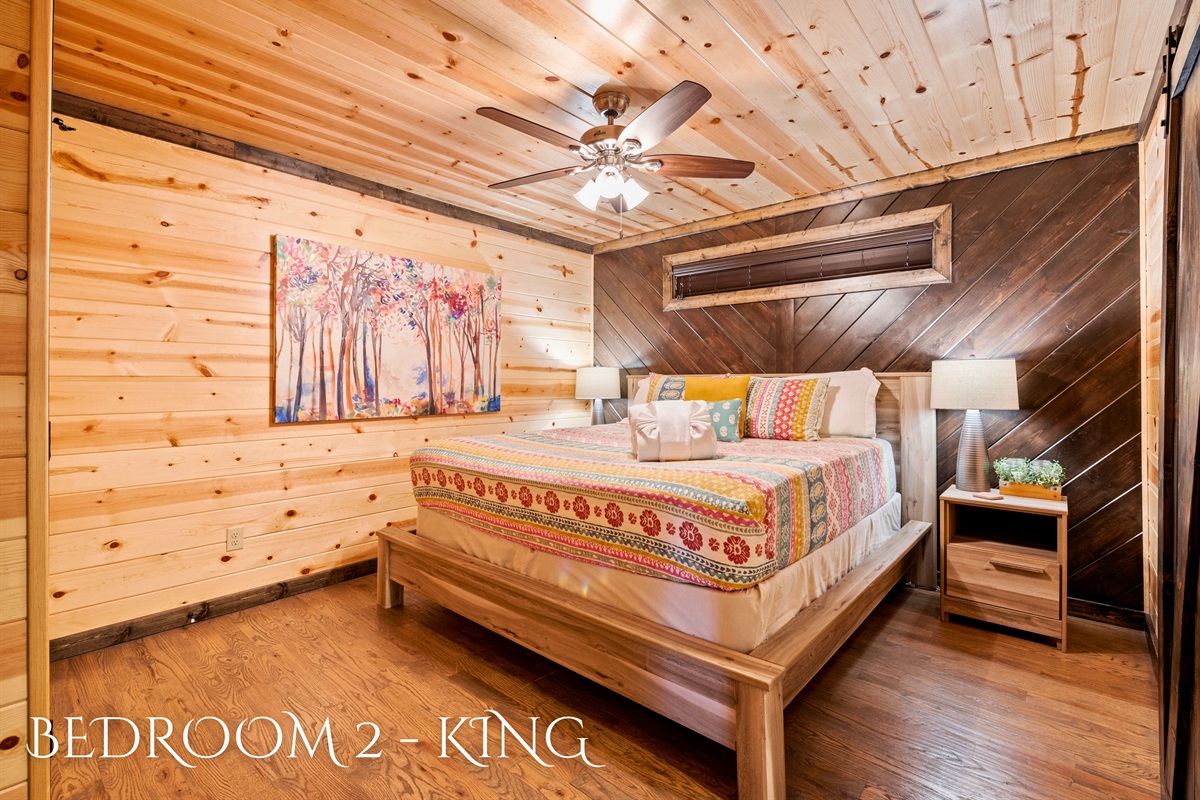 This bedroom highlights a king-sized bed with colorful bedding and a striking art piece above, blending modern style with natural wood surroundings.