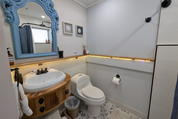 Ensuite bathroom for the queen bedroom at Villa Coral featuring a custom wooden vanity, patterned blue floor tile, an ornate blue mirror, and warm LED accent lighting in Dorado, Puerto Rico.

