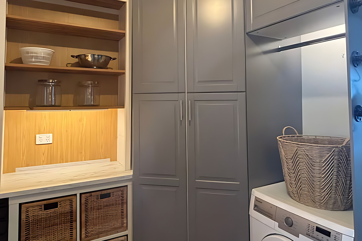 The laundry shares the scullery space, with a front-loading washing machine, a hanging rail above it, tall grey storage cupboards and open shelving fitted with wicker baskets.