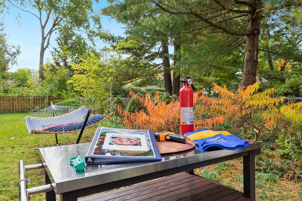 Varsity Pizza Nights are a go—our backyard prep table is stocked with gloves, fire extinguisher, infrared thermometer, lighter, and a pizza guide. Hammock and fenced yard behind complete the scene.