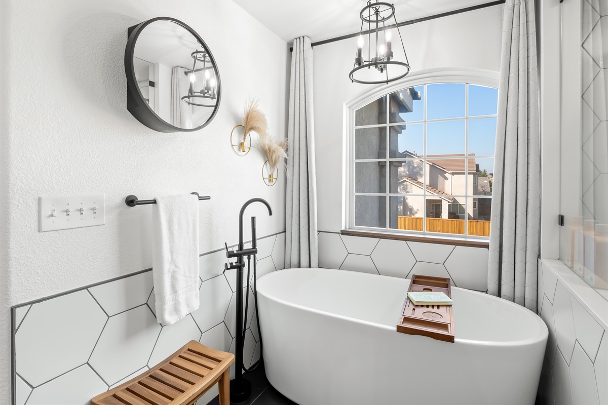 Unwind in the elegant freestanding soaking tub set beneath a large picture window, bringing in beautiful natural light. Designer lighting, modern black fixtures, and sleek tile accents create a bright, spa-inspired.