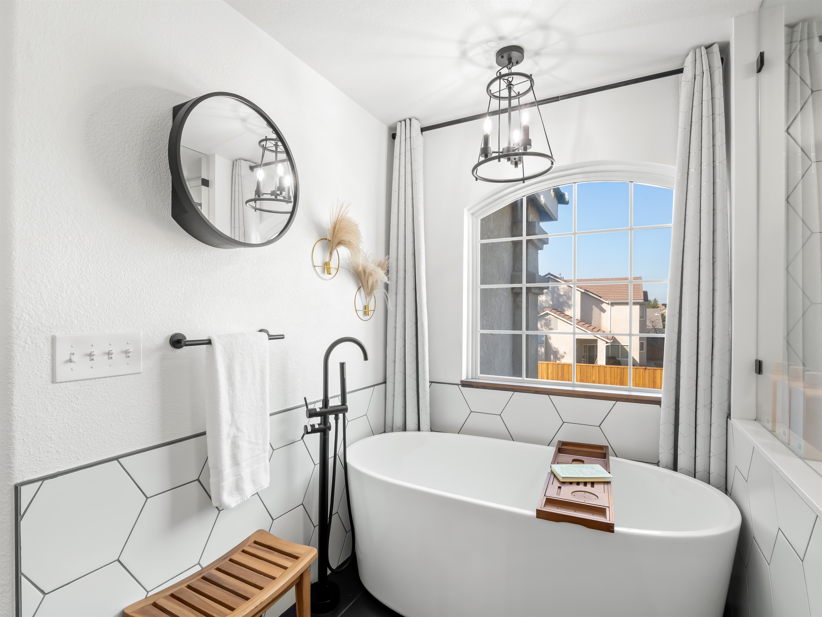 Unwind in the elegant freestanding soaking tub set beneath a large picture window, bringing in beautiful natural light. Designer lighting, modern black fixtures, and sleek tile accents create a bright, spa-inspired.