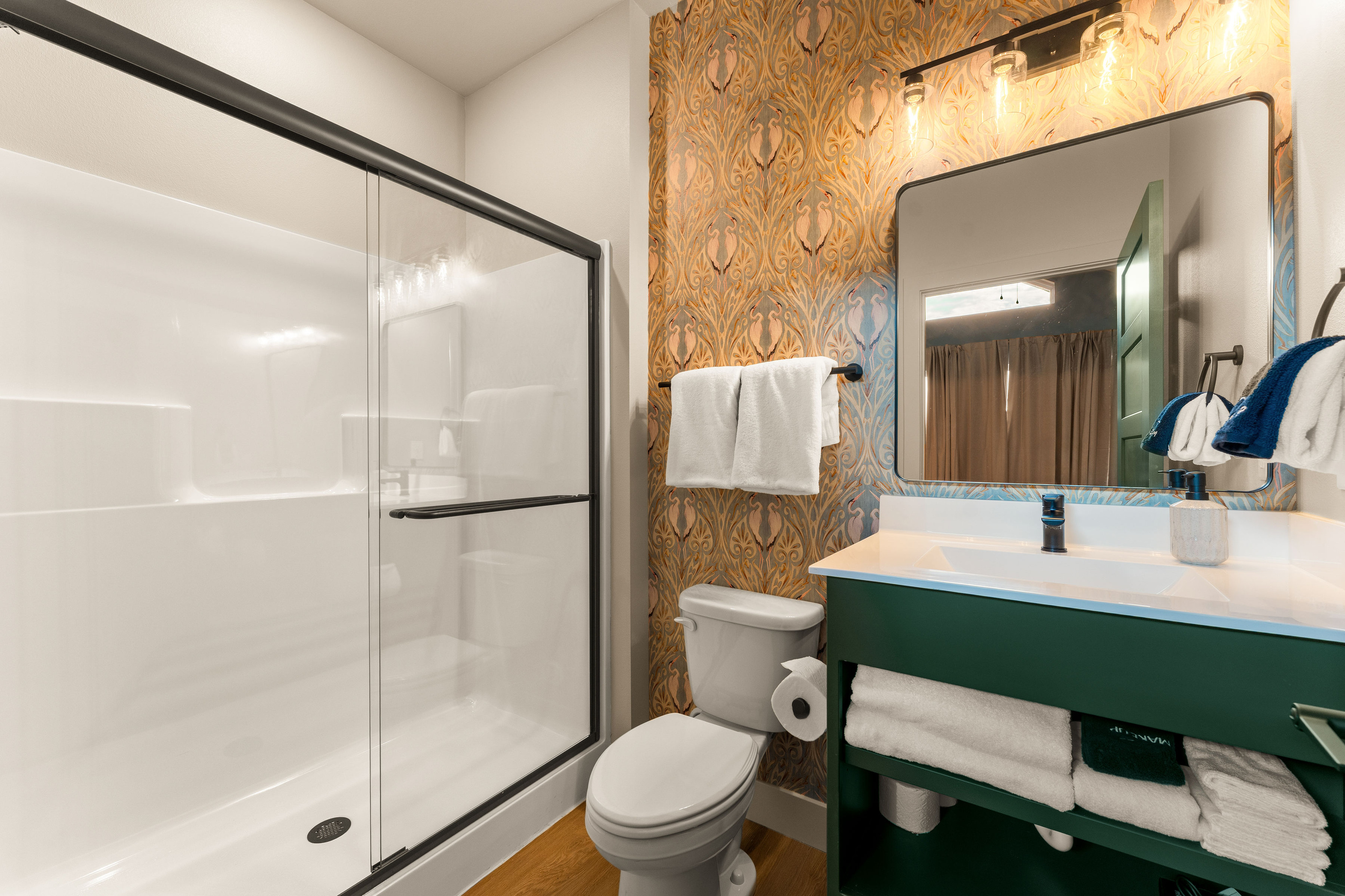 This private bathroom includes a glass-enclosed walk-in shower, rich green vanity, and intricate accent wall create a polished boutique-hotel feel.