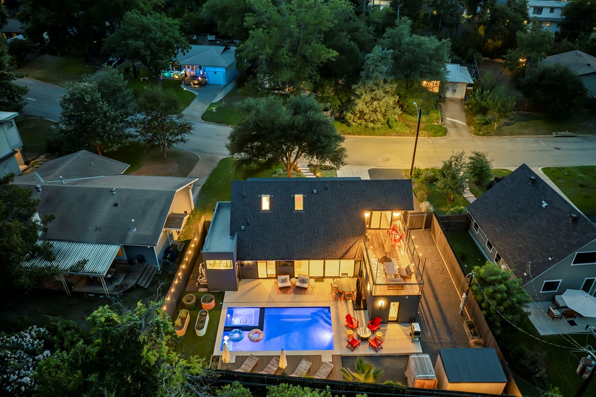 An aerial nighttime view of a modern home featuring an illuminated swimming pool with a hot tub and an inviting outdoor entertaining area with a multi-level deck, all nestled within a lush suburban neighborhood.