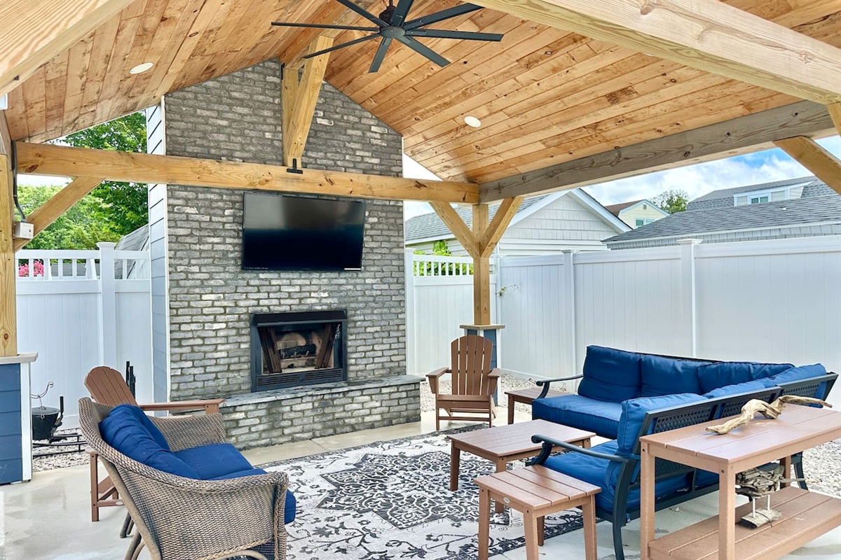 Experience luxury outdoor living in this stunning covered patio! Featuring a rustic wood ceiling, a grand stone fireplace with a TV, and plush blue seating, it’s the perfect spot for breezy summer evenings or cozy fall nights.