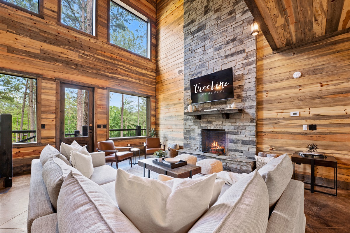 The living room is bathed in natural light, with a stone fireplace and large windows that highlight the high ceilings and wooden architecture.