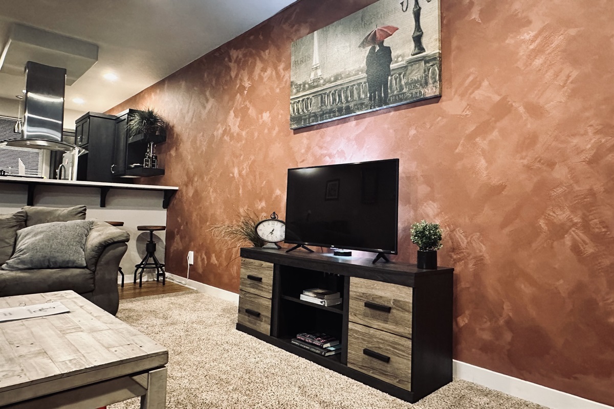 A cozy living room scene features a plush sofa with gray throw pillows, a wooden coffee table with a magazine, a modern television stand holding a flat-screen TV, a decorative clock, and a small potted plant, set against a textured rust-colored wall and visible open kitchen area.