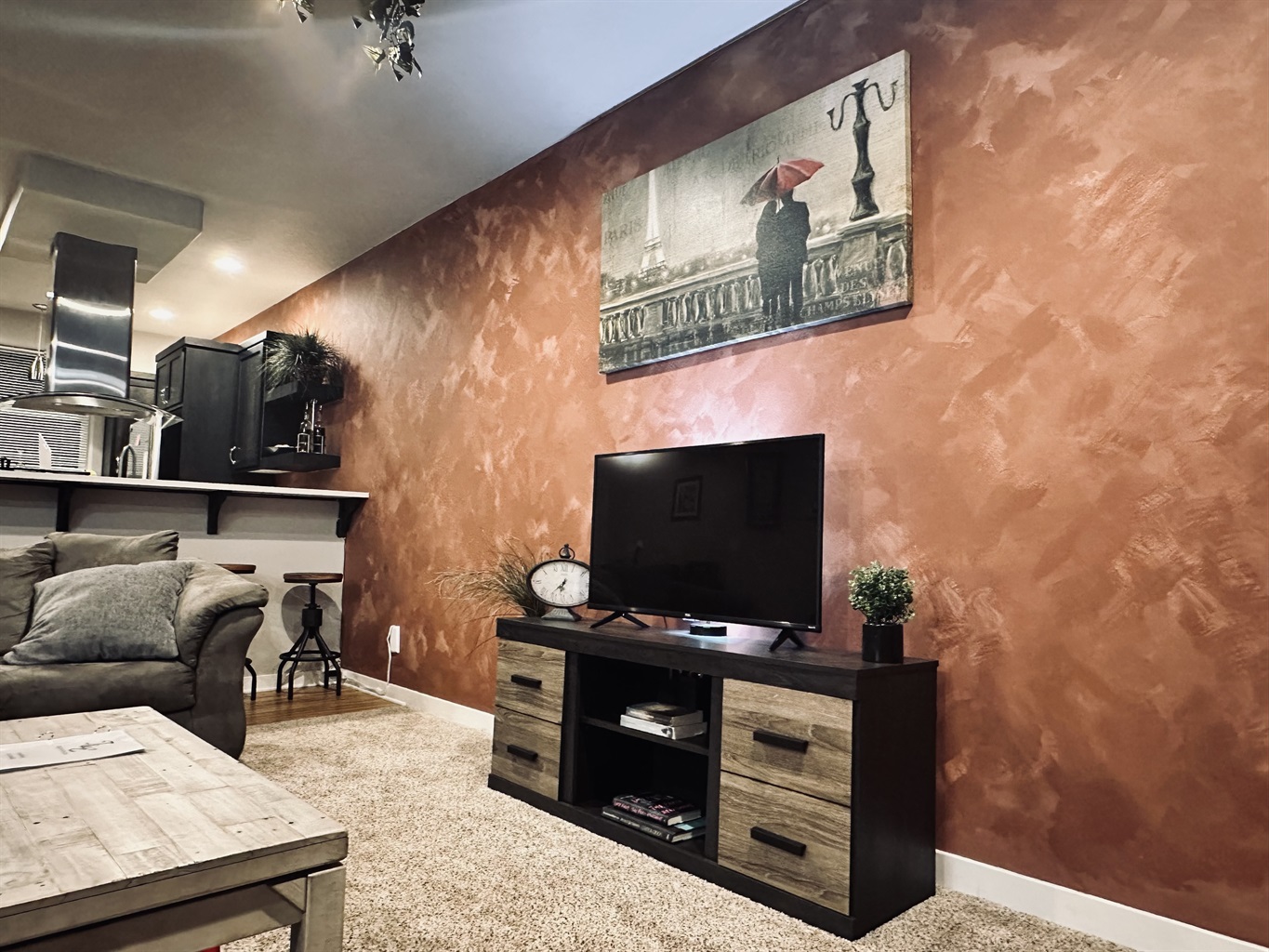 A cozy living room featuring a plush gray couch, a wooden coffee table, a contemporary black TV stand with a small plant, and a large wall art piece depicting a couple under an umbrella against a textured orange wall, with a kitchen area visible in the background.