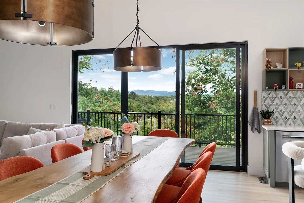 Inviting dining space with a handcrafted wood table, modern pendant lighting, and floor-to-ceiling sliding glass doors opening to a private deck, offering breathtaking mountain and treetop views for unforgettable meals
