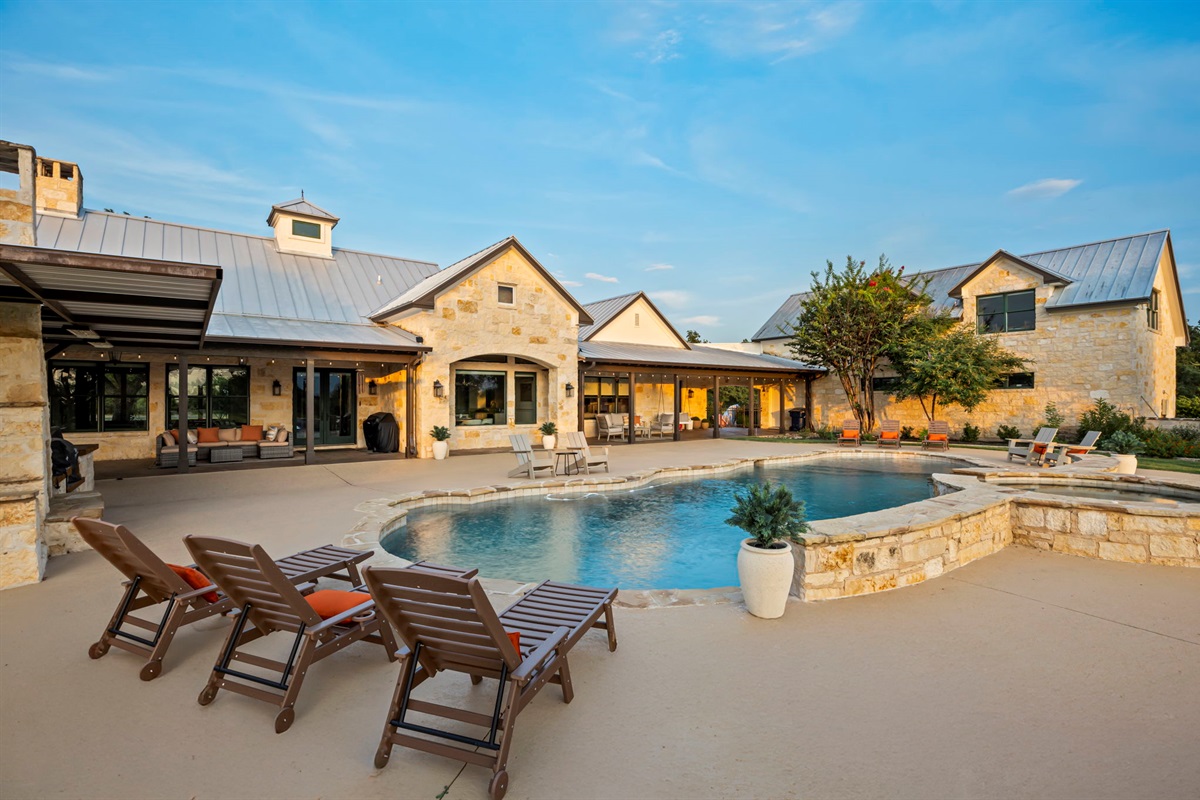 A home featuring a sprawling outdoor living area with a resort-style pool, hot tub, and ample lounging space, set against a clear blue sky.