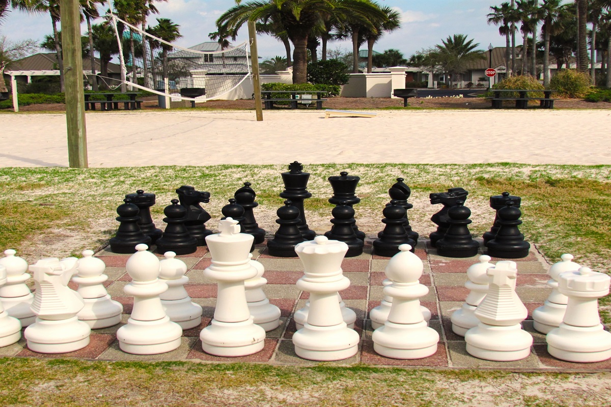 Oversized chess pieces stand ready for a match on a tiled board, situated on a grassy area adjacent to a sandy court, suggesting opportunities for both mental and physical activity while on vacation