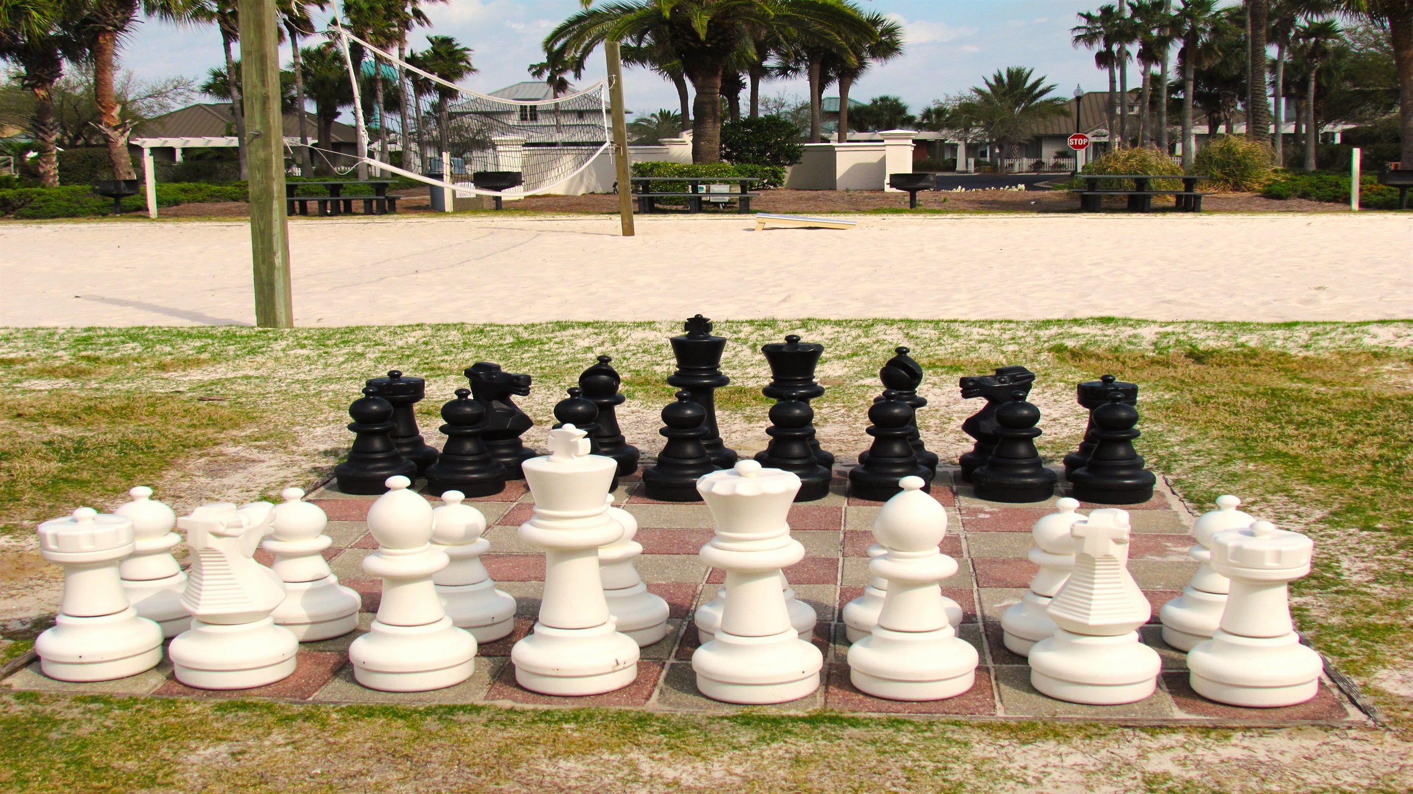 Oversized chess pieces stand ready for a match on a tiled board, situated on a grassy area adjacent to a sandy court, suggesting opportunities for both mental and physical activity while on vacation