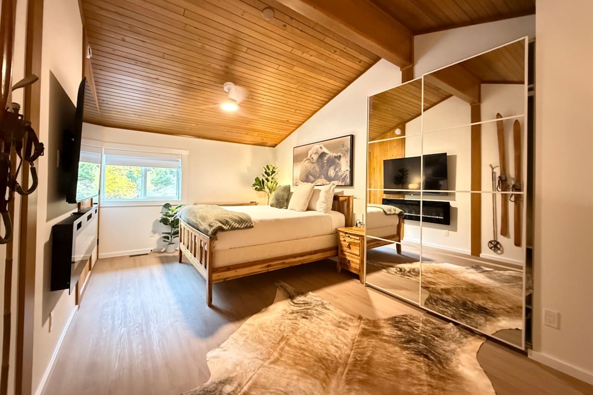 Primary Bedroom Loft Suite: A spacious loft bedroom featuring a comfortable queen-size bed, a modern built-in electric fireplace, a 65" Smart TV, closet with mirrored doors and dresser storage solutions for your gear.