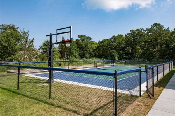 A vibrant blue-and-green outdoor court enclosed by black fencing, equipped with a regulation basketball hoop and nets for tennis or pickleball—set against a backdrop of mature trees.