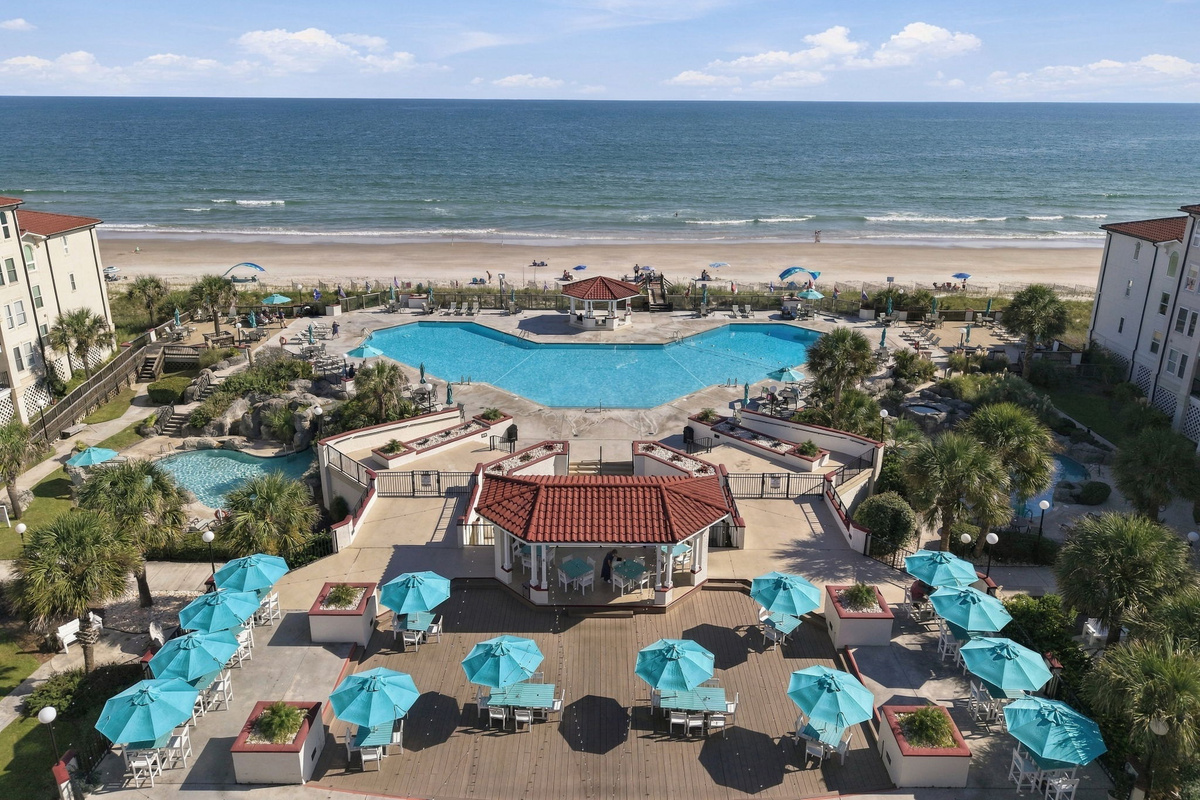 Elevated view looking over the lower dining patio with umbrella tables, central pool, poolside gazebo, and ocean shoreline.