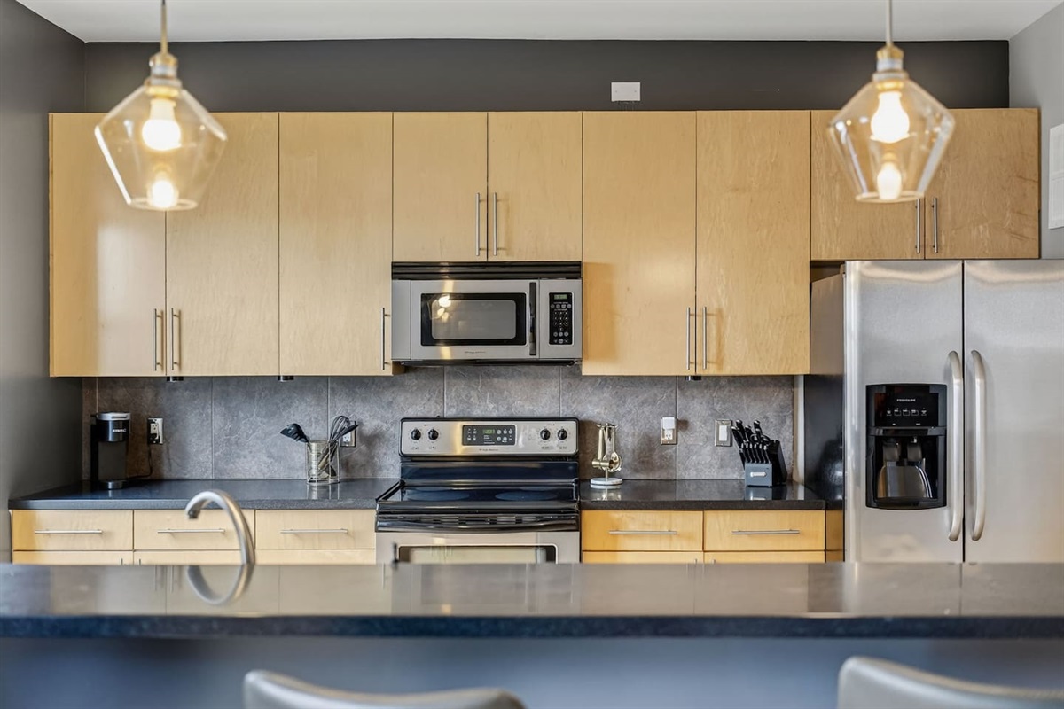Modern kitchen with sleek wood cabinetry, stainless steel appliances, and pendant lighting. Fully stocked for cooking, this stylish space is perfect for preparing meals, morning coffee, or hosting with ease.