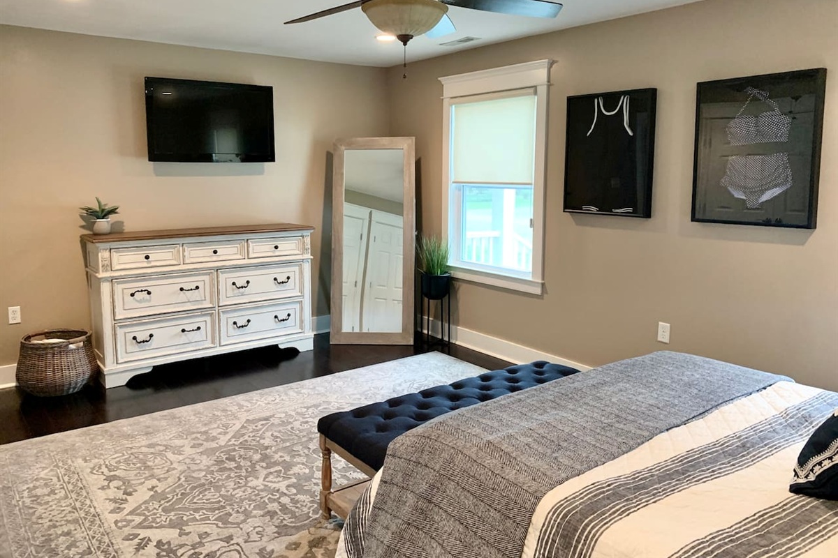 Retreat to this stylish bedroom featuring a large dresser, wall-mounted TV, and full-length mirror. Coastal-themed artwork and a plush tufted bench add a sophisticated touch, making it the perfect place to recharge after a day at the beach.
