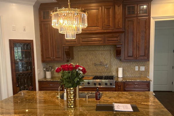 “Dream chef’s kitchen for those who love to cook and entertain, featuring rich custom wood cabinetry, oversized granite island with seating, professional-grade gas range, decorative tile backsplash, and a crystal chandelier.”