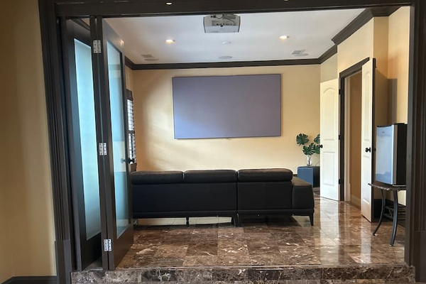 Extended master bedroom features polished marble floors, raised entry steps, dark crown molding, recessed lighting, and a private sitting area with glass doors, projector setup, and clean, modern finishes that create a luxurious, hotel-style retreat.