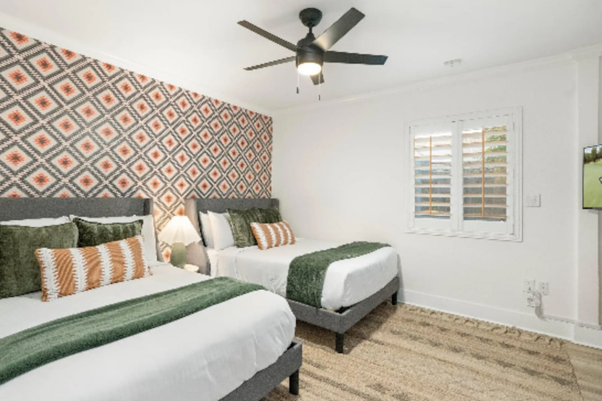 Two queen beds sit beneath a bold geometric print, with layered olive and rust pillows tying the room together. Soft natural light comes through the shutters, making this a bright, comfortable spot for friends or family sharing a room.